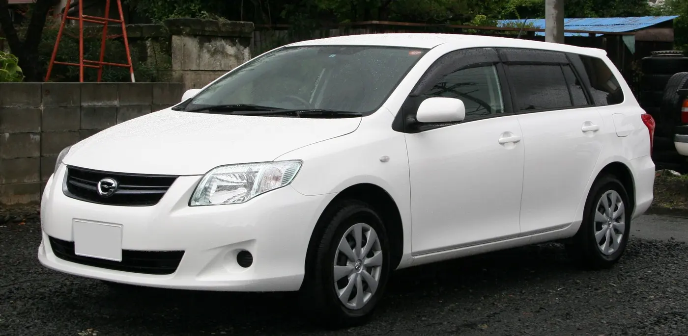A white Toyota Corolla Fielder estate parked on wet gravel beside a wall