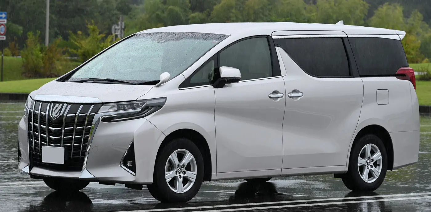 A pearl-white Toyota Alphard people carrier on a wet car park, seen from the front three-quarter angle