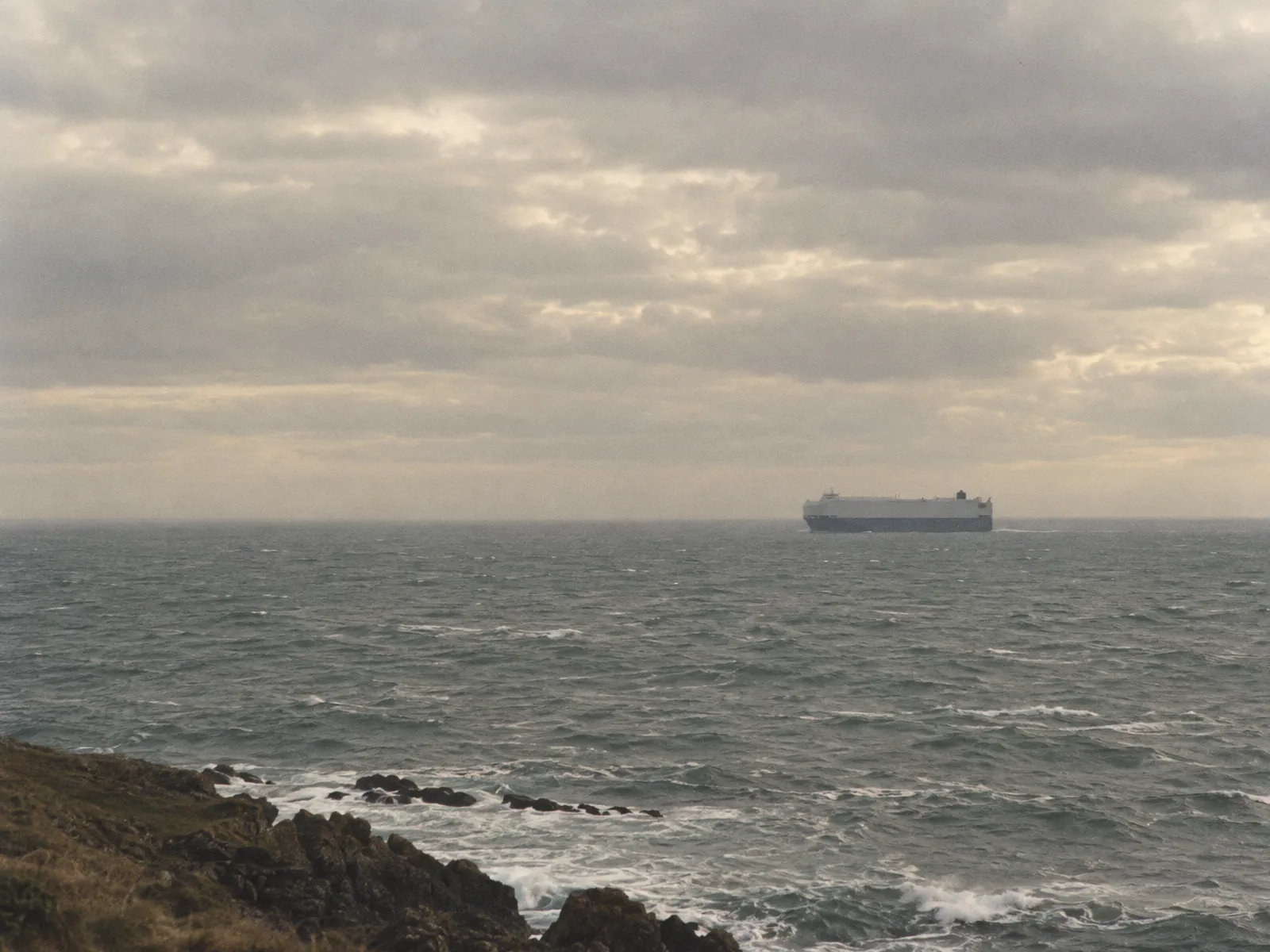 A car-carrier ship far out at sea under a heavy overcast sky, seen from a rocky headland. Illustrative image.