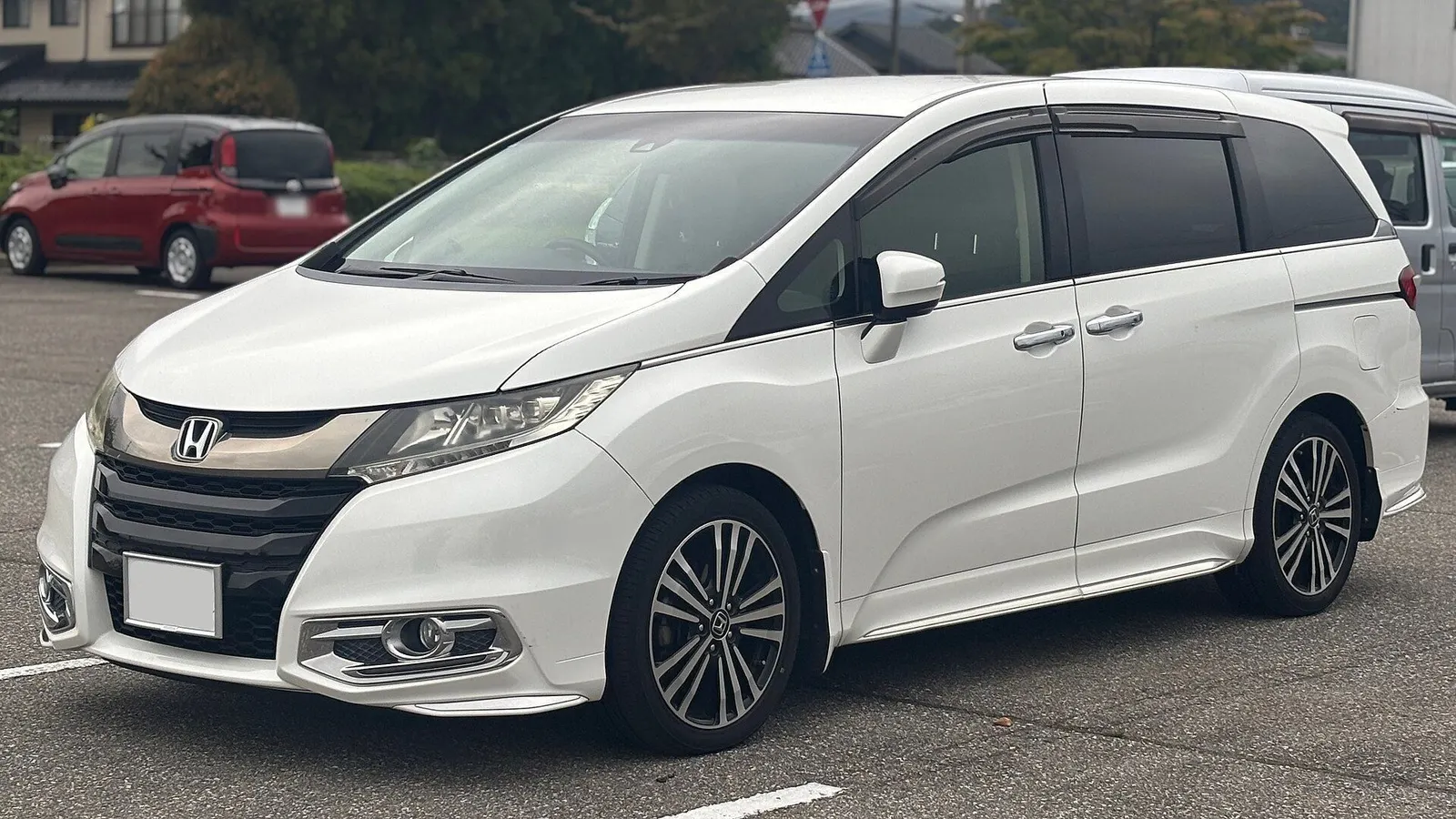 Pearl-white Honda Odyssey Absolute people carrier parked in an open car park with trees and houses behind