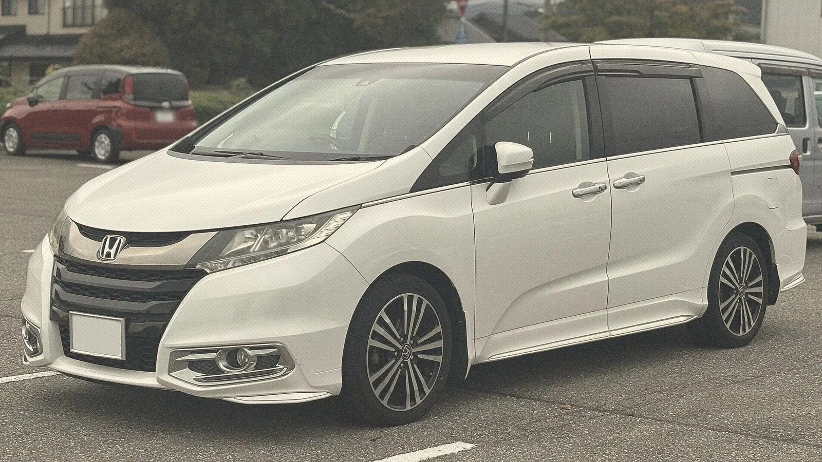 Pearl-white Honda Odyssey Absolute people carrier parked in an open car park with trees and houses behind