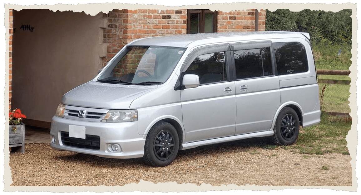 Silver Honda Step WGN Spada people carrier parked on gravel beside a red-brick barn and countryside hedge