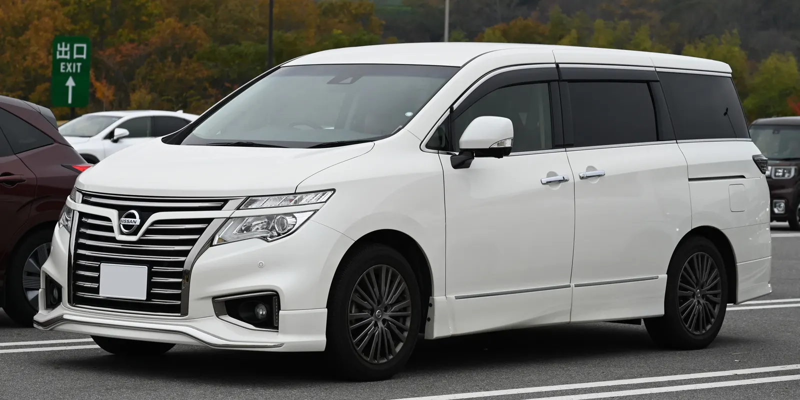 White Nissan Elgrand Highway Star people carrier in a car park with autumn woodland behind