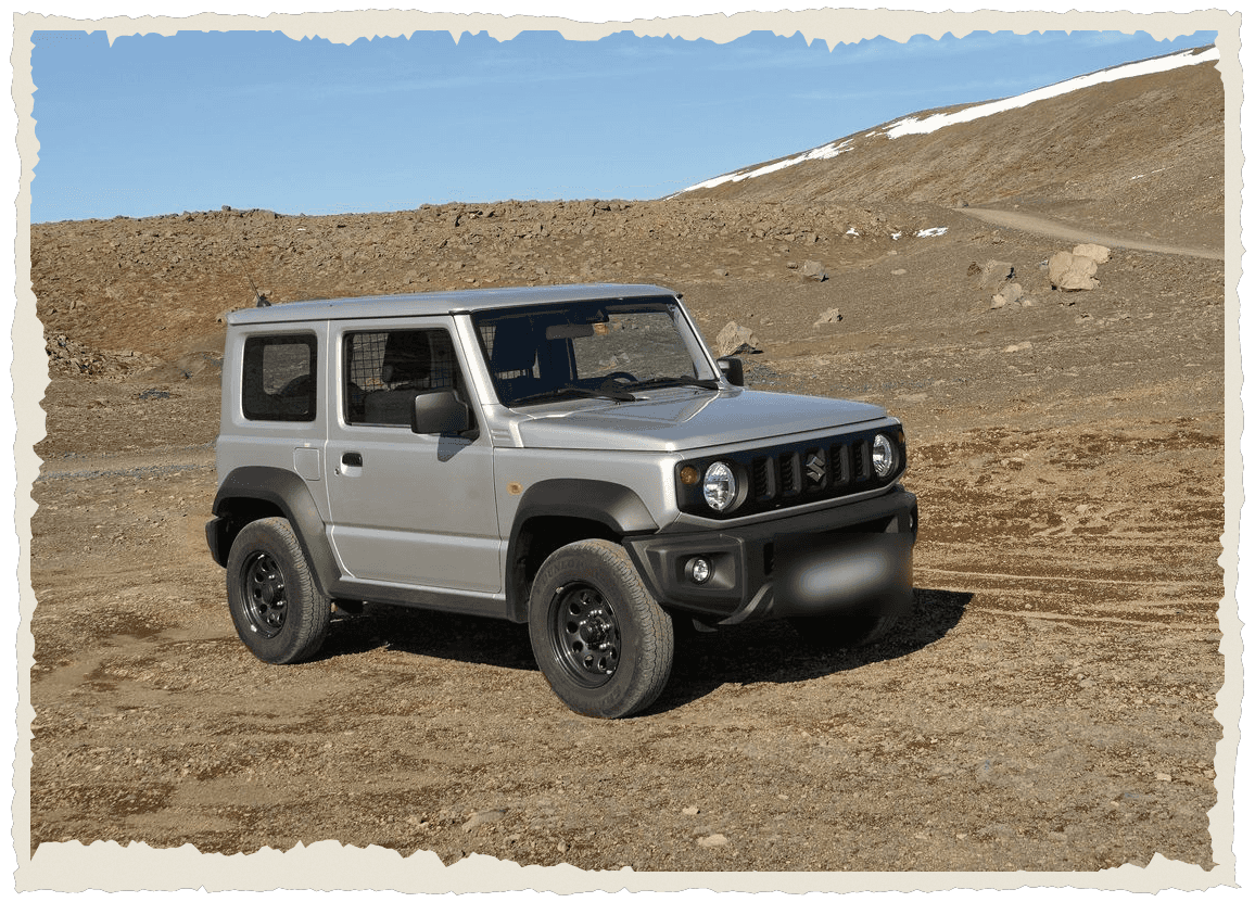 Silver Suzuki Jimny off-roader on a dirt track among bare brown hills with a snow-capped ridge and blue sky