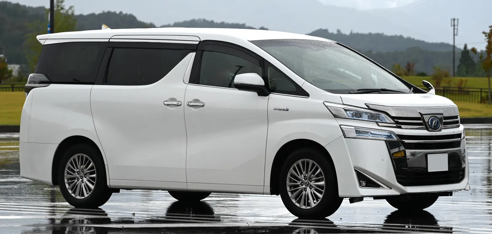 White Toyota Vellfire Hybrid people carrier on wet tarmac with green hills and low cloud behind
